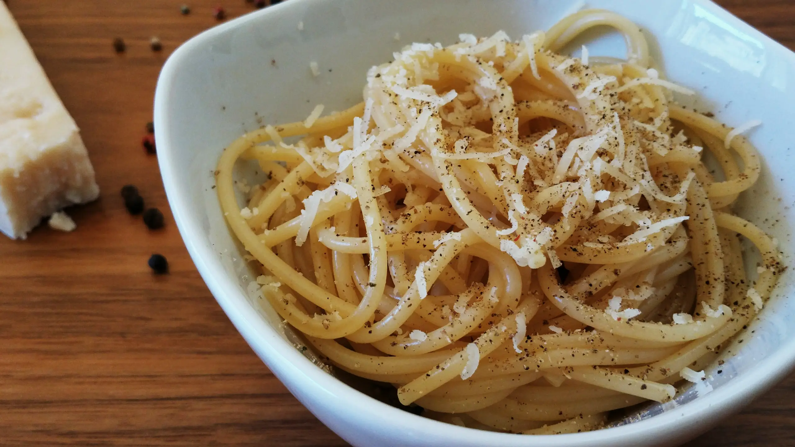 A white bowl of spaghetti cacio e pepe heaped with grated pecorino and cracked black pepper, on a wooden table.