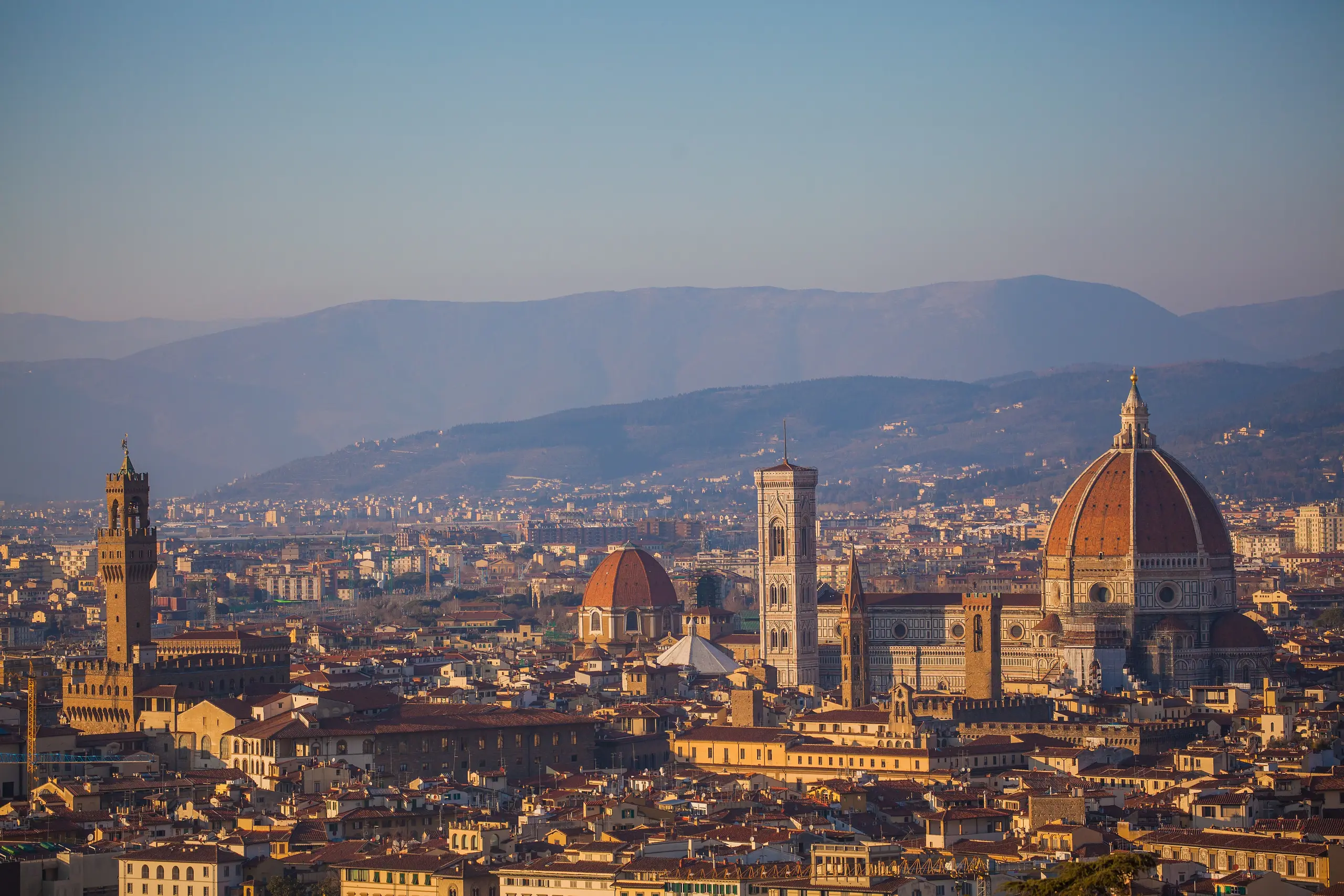 Florence's Duomo and Palazzo Vecchio tower rising over terracotta roofs at dusk, with blue hills behind.