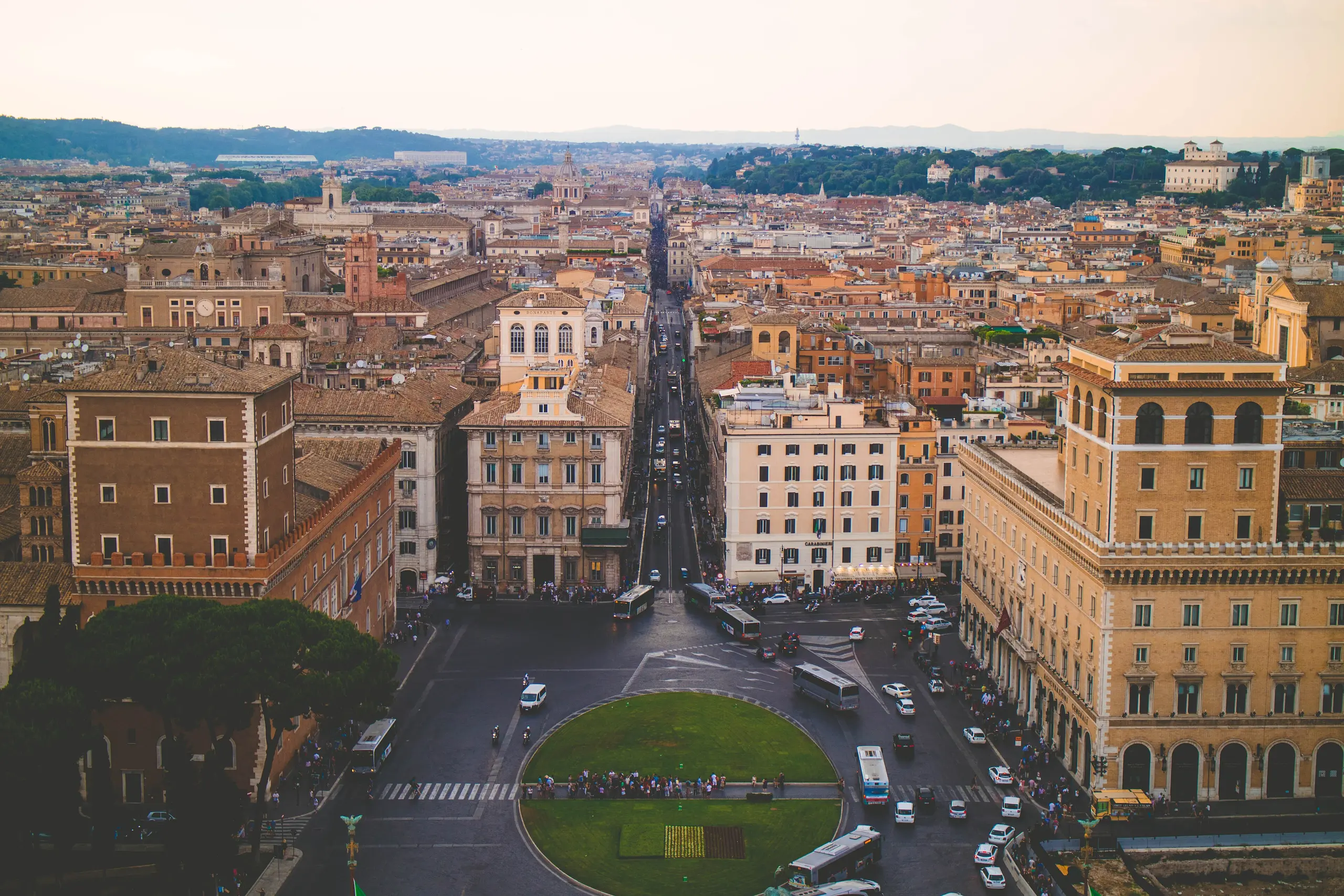 Looking down a long Roman boulevard from above at golden hour, with terracotta rooftops, umbrella pines and hills on the horizon.